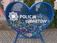 zdjęcie przedstawia serce koloru niebieskiego z białym napisem Policja, które  stoi na chodniku i wypełnione jest do połowy kolorowymi nakrętkami od butelek. Serce ma około sto osiemdziesiąt centymetrów wysokości.