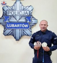 zdjęcie przedstawia Cezarego Lika lubartowskiego policjanta który brał udział w międzynarodowych zawodach w kulturystyce. Na pierwszym planie po prawej stornie zdjęcie policjant w niebieskim mundurze, w rękach trzyma  2 medale. Po lewej stronie zdjęcie logo o treści policja na białym tle na ścianie pomieszczenia.