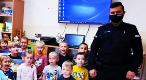 zdjęcie przedstawia na pierwszym tle dzieci a w tle policjanta który jest na spotkaniu z tymi przedszkolakami